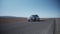 Low angle panning shot of silver small SUV car driving on road in dry landscape with mountains in distance. Morocco