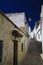 Low angle view on narrow empty alley with facades of white houses and steps upstairs contrasting with dark blue sky in traditional