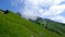 Lush green mountain hillside with jagged cliff and peaks and wispy white clouds in blue sky