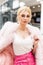 Luxury European young blonde woman in chic stylish pink fur coat in elegant white top in trendy leather skirt posing in shop