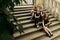 Luxury gorgeous woman in black dress posing smiling, sitting on old stairs in soft light in city, classic gothic lady outfit