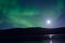 Majestic aurora borealis dancing beside full moon over mountain and calm fjord