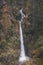Majestic vertical shot of a tall long waterfall flowing over a rocky forested cliff surface