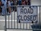 Make shift road closed sign hanging on barrier with crowd in back