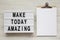 `Make today amazing` words on a light box, clipboard with blank sheet of paper on a white wooden background. From above, overhea