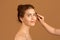 Makeup artist applying eyebrow mascara on millennial woman& x27;s face over brown studio background. Wellness and beauty