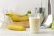 Making a milkshake. plastic disposable glass with a banana milkshake, ingredients for cooking and mixer on a light background