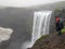 Male adult hiker adventurer hiking in raincoat back standing outdoor looking at waterfall in Laugaveur trek in stunning nature