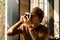 Male amateur photographer in eyeglasses focusing to make photos out of window sitting on windowsill.Young bearded man resting in