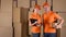 Male anf female couriers in orange uniform standing against brown cardboard boxes backround. Delivery company staff, 4K