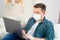 Male blogger in medical mask working on couch