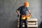 A male builder or repairman puts on a tool belt. The constructor stands against a gray wall wearing an orange hard hat