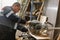 A male carpenter in uniform, holds a grip on a lathe with a sharp round knife slicing a wood bar.