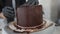 Male Caucasian hands in black gloves spinning rotating table adding chocolate topping on delicious baked cake