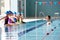 Male Coach In Water Gives Children Instructions In Swimming Lesson As They Sit On Edge Of Pool