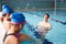 Male Coach In Water Gives Children Instructions In Swimming Lesson As They Sit On Edge Of Pool