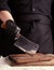 Male cook in black uniform and black latex gloves holds a large sharp meat knife over a cutting board