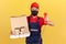 Male courier in blue overalls red t-shirt and cap with protection mask holding landline phone and coffee with pizza, call to order