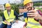 male engineers in safety vests and helmets with blueprints taking selfie on smartphone