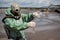 A male environmentalist in a green protective suit and gas mask takes a sample of water. The scientist is doing a