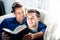 Male gay couple cuddling and reading together on large couch next to patio doors with bright sunlight shining on them