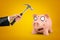 Male hand holding hammer over pink cartoon face piggy bank on yellow background