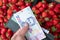 Male hand holding hryvnia banknotes above fresh strawberries in boxes