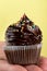 Male hand holding or presenting tasty appetizing chocolate cupcake. Yellow Background. Closeup.