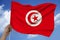 Male hand holds against the sky with clouds the national flag of Tunisia on a luxurious texture of satin, silk with waves, close