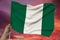 Male hand holds a colored Nigeria flag on a background of the sky with clouds on a luxurious satin texture, silk with waves,