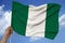 Male hand holds a colored Nigeria flag on a background of the sky with clouds on a luxurious satin texture, silk with waves,