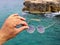 Male hand holds sunglasses with reflexions of cloudy sky in front of stony coast above the sea