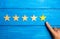 The male hand points to the fifth yellow star on a blue wooden background. Five Stars. Rating of restaurant or hotel, application.