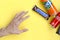 Male hand reaches for Snickers chocolate bar in brown wrapping, red Coca Cola and orange Fanta tin can on yellow pastel background