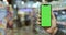 Male hand showing modern black smartphone with tamplate green screen in vertical position at blurred supermarket