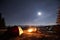 Male hiker have a rest in his camp near the forest at night under beautiful night sky full of stars and the moon