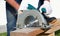 A male joiner works with a hand-held circular saw. A builder is sawing a board at the construction site of a new house