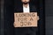 Male office worker in suit holding cardboard sign written looking for a job text, standing outside business center