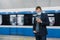 Male passenger wears face mask poses at platform, waits for train, commutes by underground, concentrated in smartphone device,