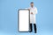 Male physician in white uniform pointing finger at huge cellphone with blank screen on blue studio background, mockup