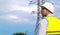 A male power engineer in a white helmet checks a power line using data from electrical sensors on a tablet. High voltage