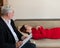 A male psychotherapist writes on a clipboard during a patient consultation. An emotional woman in a red dress lies on a