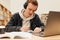 Male student preparing exams at desk in library. Smiling guy in headphones writing on notebook