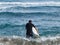 Male in swimsuit going in water with surfboard.