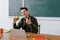 Male teacher in glasses looking at camera, holding apple and showing thumb up sign at computer desk