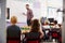 Male Teacher Standing At Whiteboard Teaching Maths Lesson To Elementary Pupils In School Classroom