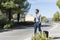 A male traveler with a suitcase and an empty cardboard sign stands on the road. A guy with glasses and a beard. Free hitchhiking.