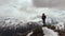 A male traveller with a backpack stands on top of a cliff against the backdrop of snow-capped mountain peaks and low