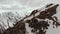 A male traveller with a backpack stands on top of a cliff against the backdrop of snow-capped mountain peaks and low