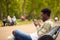 Male Vlogger Or Social Influencer Sitting On Bench In City Park Looking At Mobile Phone